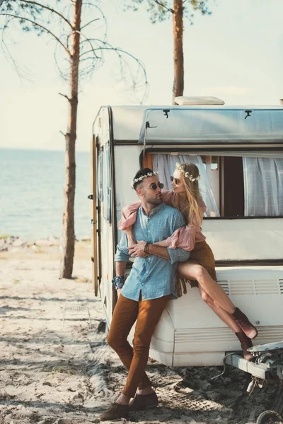 Pareja hippie en coronas y gafas de sol abrazando y sentado en el remolque - foto de stock