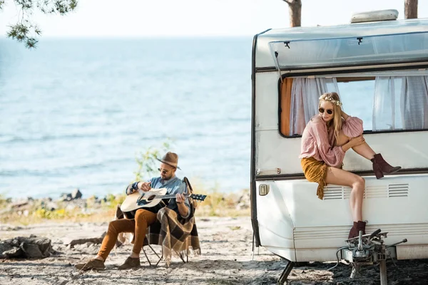 Hermosa chica hippie sentado en autocaravana mientras que el hombre tocando la guitarra cerca del mar - foto de stock