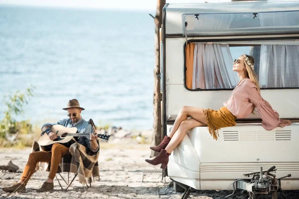 Attractive girlfriend sitting on trailer while man playing guitar near the sea — Stock Photo