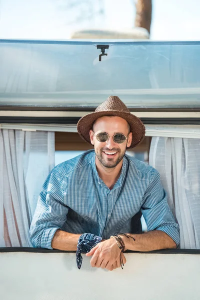 Cheerful man in sunglasses and hat looking from window of campervan — Stock Photo