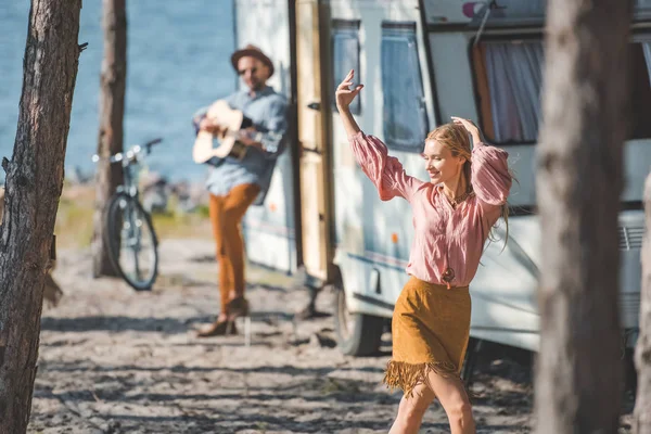 Jovem hippie mulher dançando enquanto homem tocando guitarra perto trailer — Fotografia de Stock