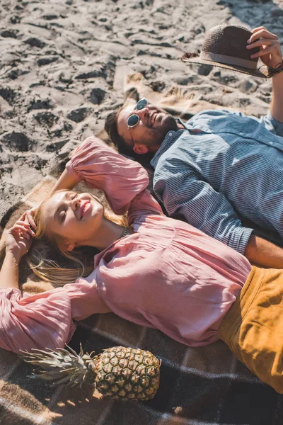 Пара хиппи с ананасом и шляпой расслабляющие на одеяло на песке — стоковое фото