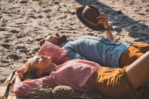 Beautiful couple lying on blanket with pineapple on sand — Stock Photo