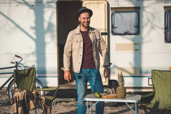 Hombre feliz de pie cerca de la mesa con piña y aguacate en el campamento - foto de stock