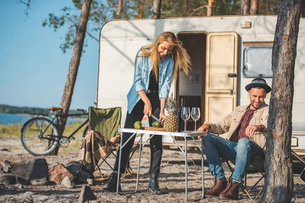 Hermosa chica cortar aguacates mientras feliz hombre descansando cerca en campamento - foto de stock