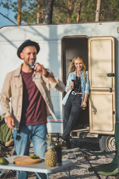 Messa a fuoco selettiva di bella donna in piedi al camper con un bicchiere di vino guardando fidanzato — Foto stock