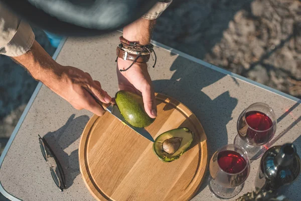 Vista recortada del hombre cortando aguacate sobre tabla de madera sobre mesa con vino - foto de stock