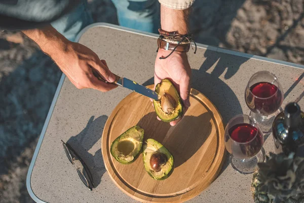 Partial view of man cutting avocado on wooden board on table with wineglasses and sunglasses — Stock Photo