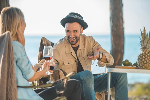 Улыбающаяся пара с бокалами вина разговаривает во время пикника у моря — стоковое фото