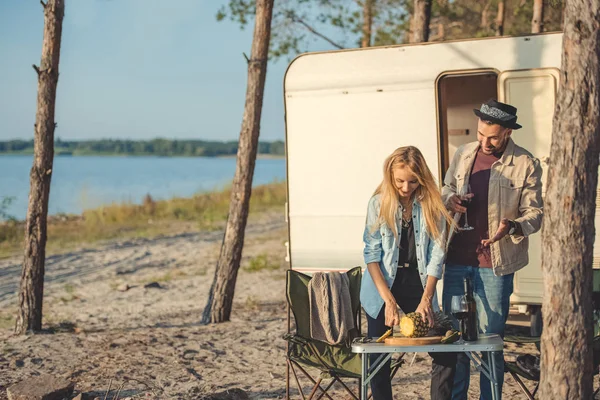 Young woman cutting pineapple while man with glass of wine talking to her near trailer — Stock Photo