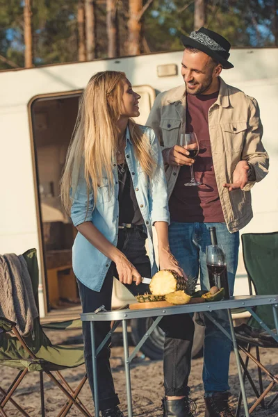 Beautiful girl cutting pineapple and looking man with wine near campervan — Stock Photo