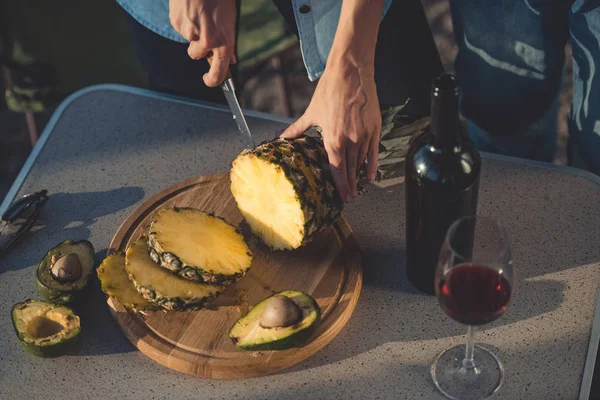 Cropped view of woman cutting pineapple and avocados on wooden board near bottle of red wine — Stock Photo