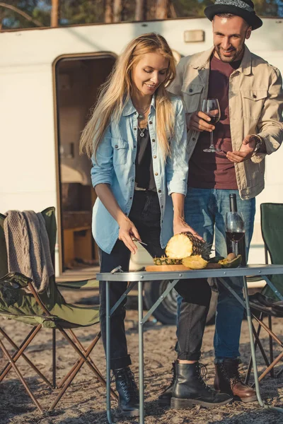 Schönes Mädchen schneidet Ananas, während Mann mit Wein in der Nähe von Wohnmobil mit ihr spricht — Stockfoto