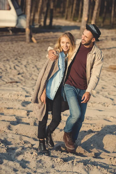 Улыбающийся мужчина и привлекательная женщина обнимаются и гуляют по песчаному пляжу — стоковое фото