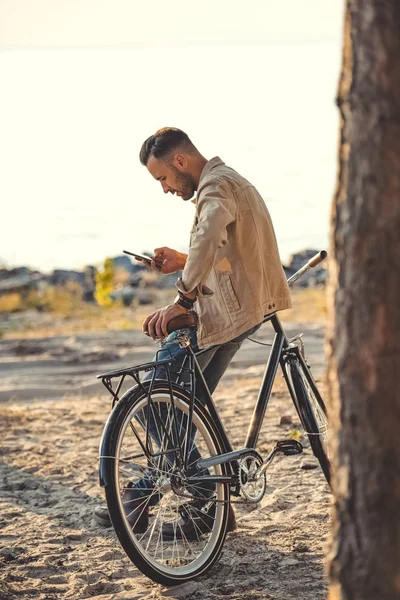 Joven hombre usando gadget y apoyándose en bicicleta cerca del mar - foto de stock