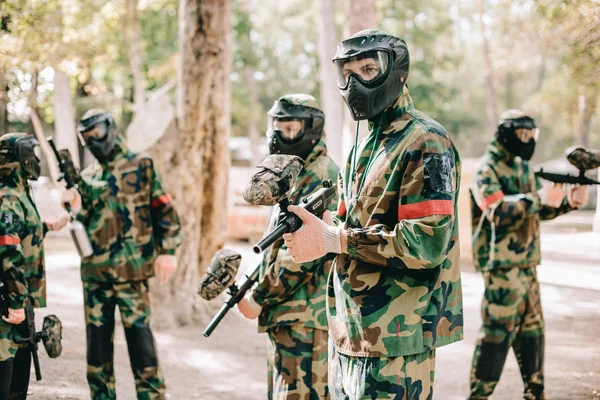 Equipo de paintball en uniforme y máscaras protectoras jugando paintball con pistolas marcadoras al aire libre - foto de stock