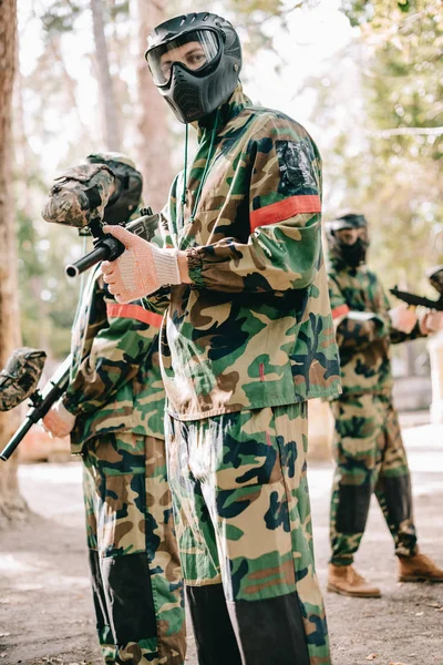 Male paintball player in uniform and goggle mask looking at camera and standing with his team outdoors — Stock Photo