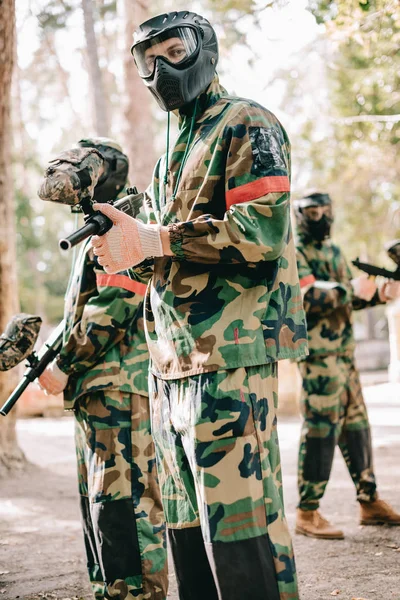 Jugador de paintball masculino en uniforme y máscara de gafas mirando a la cámara y de pie con su equipo al aire libre - foto de stock