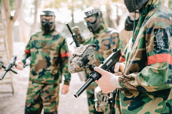 Partial view of paintball team in camouflage and protective masks standing with paintball guns outdoors — Stock Photo