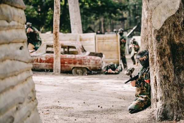 Female paintball player in goggle mask and camouflage sitting with paintball gun while her team sitting behind near broken car outdoors — Stock Photo