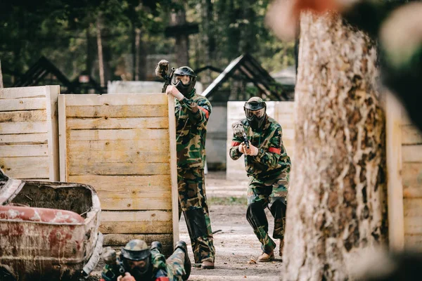 Selective focus of paintball team in uniform and protective masks playing paintball with marker guns outdoors — Stock Photo