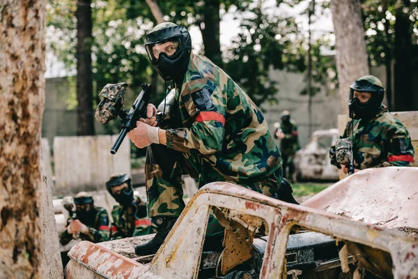 Jugador de paintball masculino con pistola marcadora sentado en el coche roto mientras su equipo de pie detrás al aire libre - foto de stock