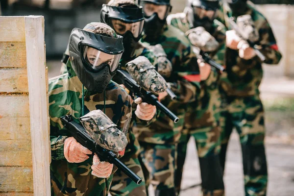 Foyer sélectif de la joueuse de paintball tenant le pistolet marqueur avec son équipe dans des masques de protection et de camouflage jouant au paintball à l'extérieur — Photo de stock