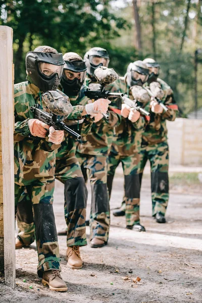 Female paintball player holding marker gun with her team in protective masks and camouflage playing paintball outdoors — Stock Photo