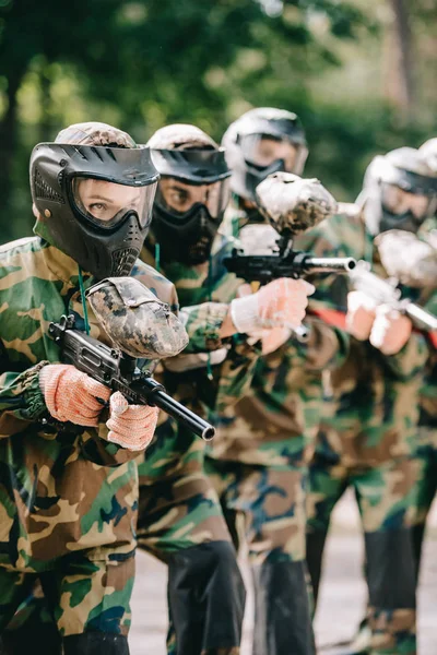 Joueuse de paintball concentrée tenant un pistolet marqueur avec son équipe dans des masques de protection et de camouflage jouant au paintball à l'extérieur — Photo de stock