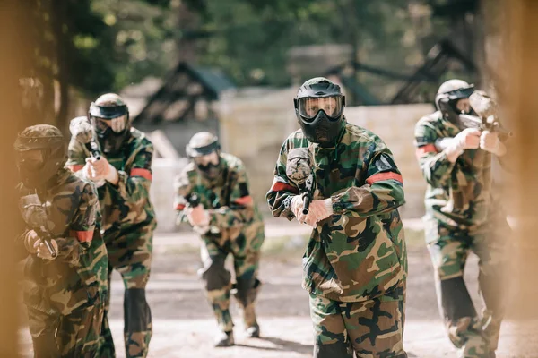 Selective focus of paintball team in uniform and protective masks playing paintball with marker guns outdoors — Stock Photo