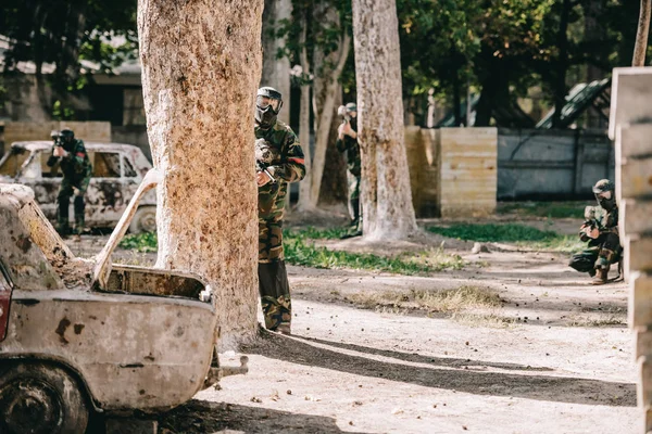 Paintball team in uniform and protective masks playing paintball with marker guns near broken car outdoors — Stock Photo