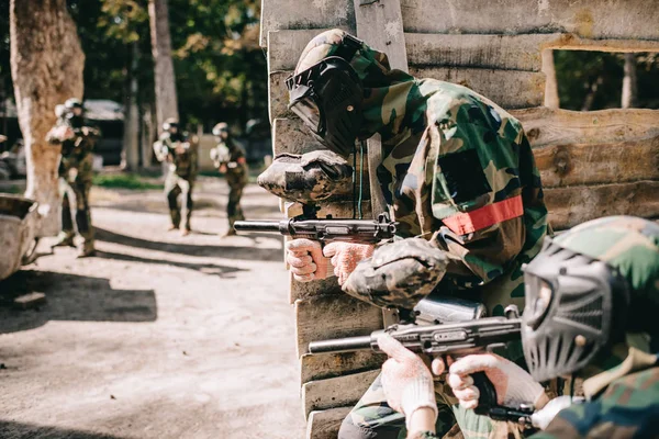 Selective focus of paintball player in protective mask holding marker gun and his teammate hiding behind wooden wall outdoors — Stock Photo