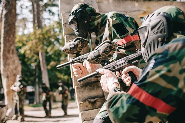 Foco seletivo do jogador de paintball em máscara protetora segurando arma marcador e seu companheiro de equipe se escondendo atrás da parede de madeira ao ar livre — Fotografia de Stock