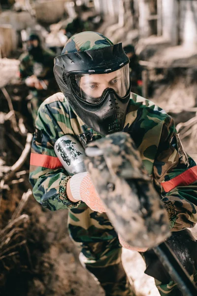 Selective focus of male paintball player in goggle mask and camouflage aiming by paintball gun from ditch outdoors — Stock Photo