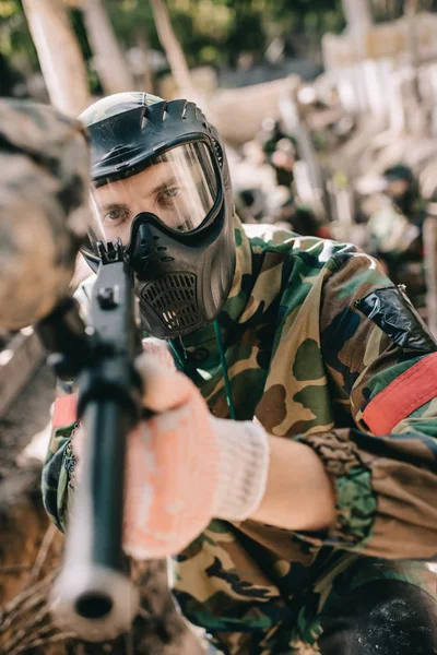 Selective focus of male paintball player in goggle mask and camouflage aiming by paintball gun outdoors — Stock Photo