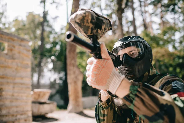 Concentrated male paintball player in goggle mask and camouflage aiming by paintball gun outdoors — Stock Photo