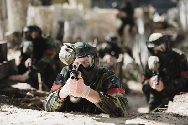 Selektiver Fokus des konzentrierten Paintballspielers in Schutzmaske, der mit der Markierungspistole und seinem Team auf den Hintergrund im Freien zielt — Stockfoto