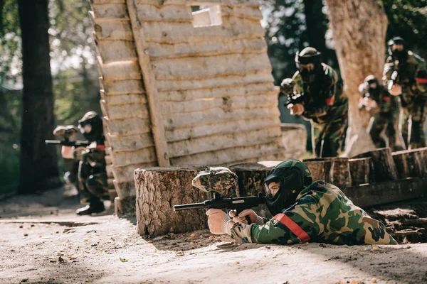 Joueur de paintball étendu sur le sol et visant par pistolet marqueur tandis que son équipe se cache derrière un mur en bois à l'extérieur — Photo de stock