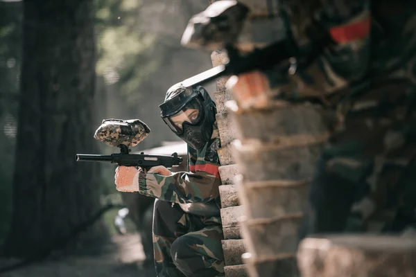 Selective focus of paintball player in protective mask holding marker gun and his teammate hiding behind wooden wall outdoors — Stock Photo