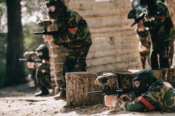 Jogador de paintball focado deitado no chão e apontando com arma de marcador, enquanto sua equipe se esconde atrás da parede de madeira ao ar livre — Fotografia de Stock