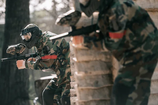 Селективный фокус пейнтболиста в защитной маске, держащего маркерную пушку, и его товарища по команде, прячущегося за деревянной стеной — стоковое фото