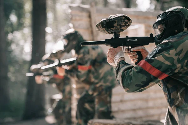 Selective focus of paintball player in goggle mask aiming with marker gun and his team on background outdoors — Stock Photo