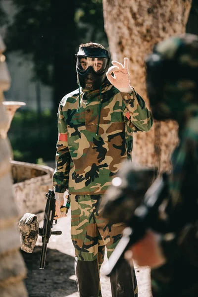 Jugador de paintball masculino en camuflaje y máscara de gafas mostrando signo ok a su equipo al aire libre - foto de stock