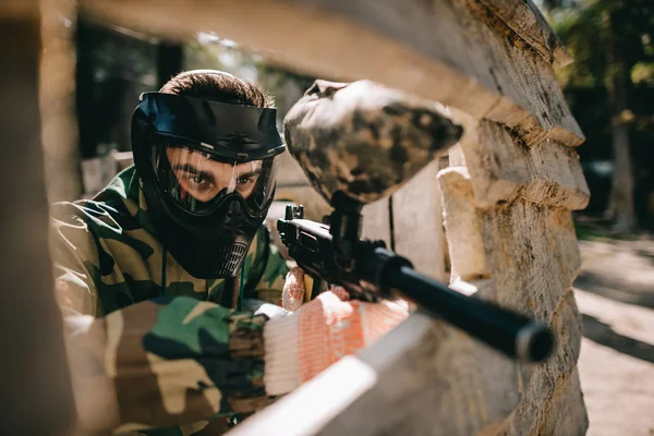 Concentrated male paintball player in goggle mask and camouflage aiming by paintball gun outdoors — Stock Photo