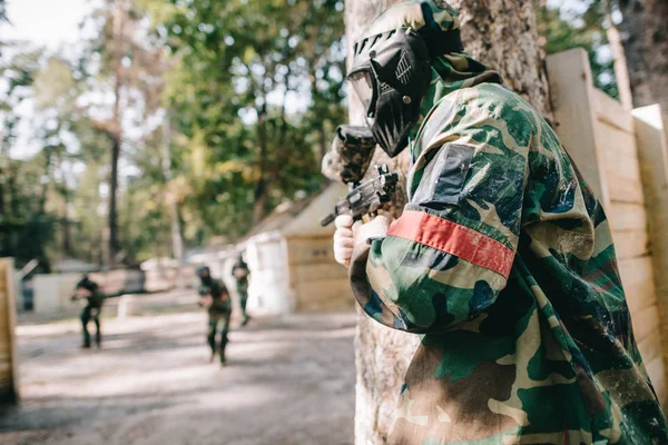 Male paintballer in camouflage and protective mask holding marker gun and hiding behind tree while other team running behind outdoors — Stock Photo