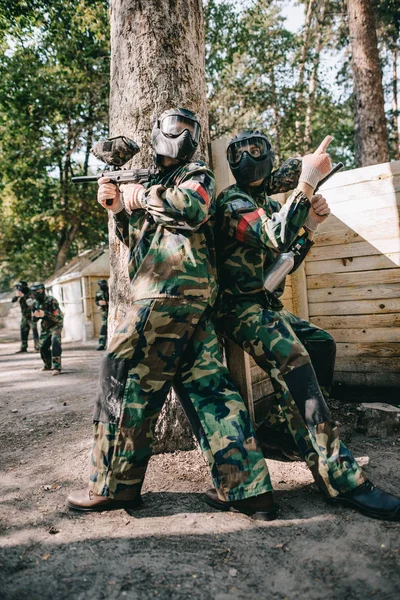 Male paintball player in goggle mask and camouflage holding marker gun and pointing by finger to teammate near tree outdoors — Stock Photo