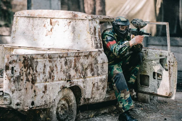 Male paintball player in goggle mask and camouflage aiming by paintball gun from broken car outdoors — Stock Photo