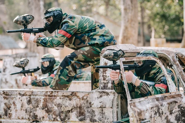 Paintball team in protective masks and camouflage aiming by marker guns from broken car outdoors — Stock Photo