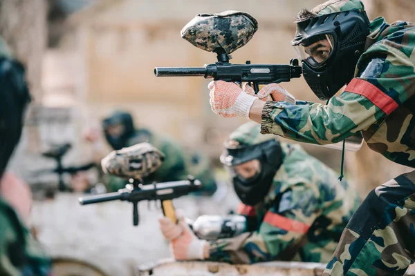 Enfoque selectivo del jugador de paintball en máscara protectora apuntando con pistola marcadora y su compañero de equipo detrás al aire libre - foto de stock