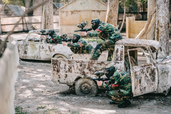 Equipo de paintball en camuflaje y máscaras de gafas apuntando con pistolas marcadoras de coches rotos al aire libre - foto de stock